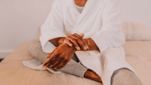 Black person wearing a white robe rubbing lotion onto their arms.