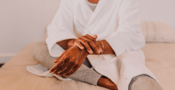 Black person wearing a white robe rubbing lotion onto their arms.