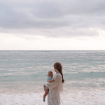 A white woman carrying a baby looks out onto an overcast horizon over the ocean.