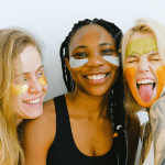 Three young women, one black and two white, smile against a white background. They are wearing neutral colours and summer skincare products on their faces.