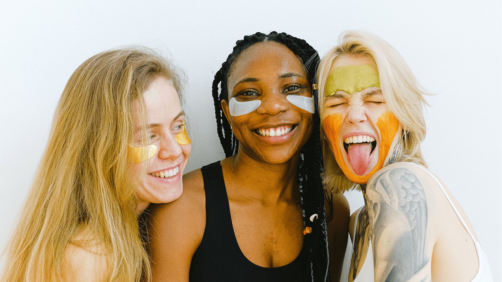 Three young women, one black and two white, smile against a white background. They are wearing neutral colours and summer skincare products on their faces.