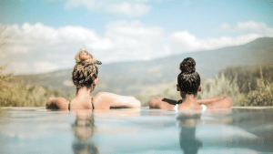 Two young women are swimming in an infinity pool, looking out onto a panoramic view.