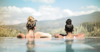 Two young women are swimming in an infinity pool, looking out onto a panoramic view.